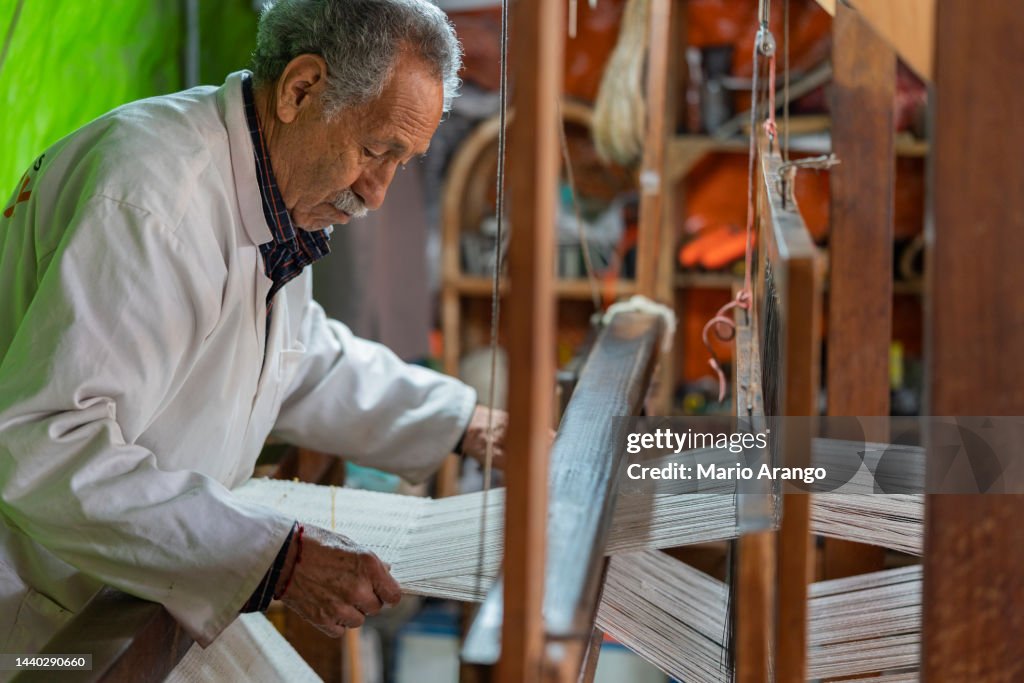 An older man, an artisan by profession, is in his factory where he weaves handcrafted looms.