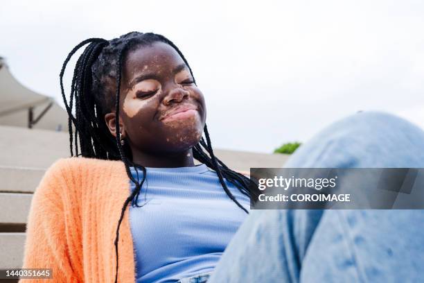 young african woman sitting on some stairs with a smile and her eyes closed.young woman with vitiligo. - one person smile outdoors stock-fotos und bilder