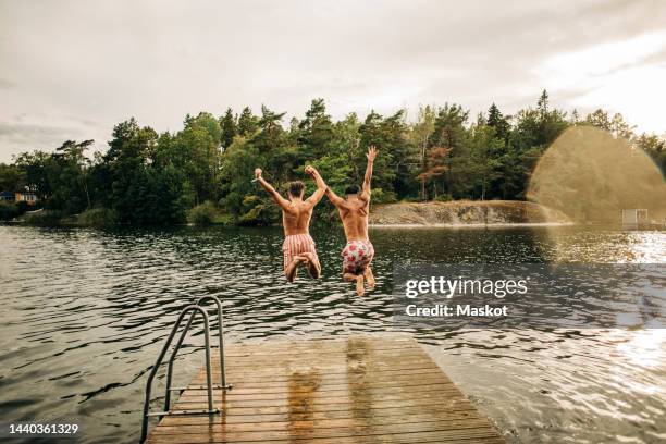carefree male friends with arms raised jumping in lake during vacation - jetty stock pictures, royalty-free photos & images