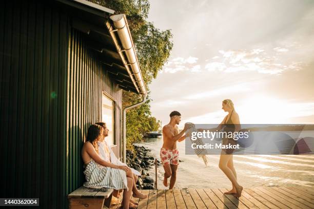 woman passing towel to male friend while spending vacation at lake during sunset - standing water stockfoto's en -beelden