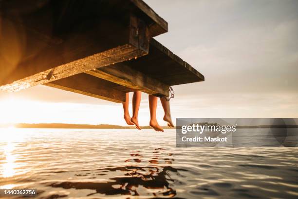 low section of female friends sitting on jetty by lake during sunset - stockholm summer stock-fotos und bilder