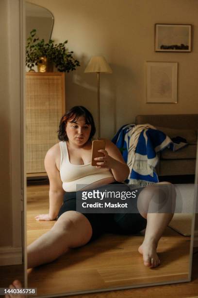 full length of young voluptuous woman taking selfie through smart phone while sitting against mirror at home - chubby foto e immagini stock