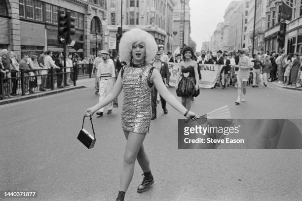 Cross-dressing marcher wearing a multi-coloured, sequinned mini-dress with handbag and fan accessories, ahead of a banner promoting the LGBT rights...