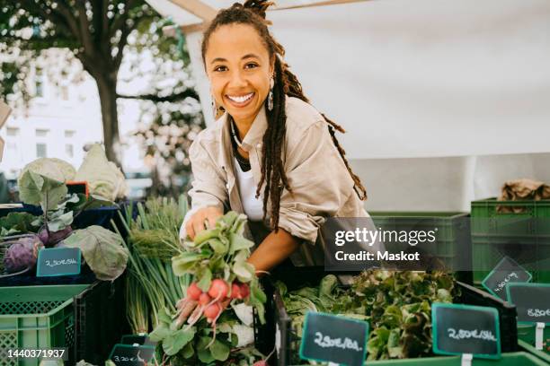 portrait of happy female owner holding vegetable near crates in stall at market - marktstand stock-fotos und bilder