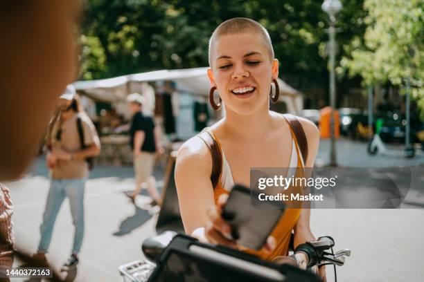 smiling woman with shaved head paying via tap to pay while shopping at flea market - brand name mobile payment foto e immagini stock