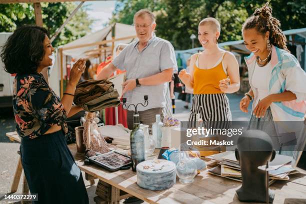 side view of happy female owner holding dress with customers shopping near stall at flea market - rommelmarkt stockfoto's en -beelden