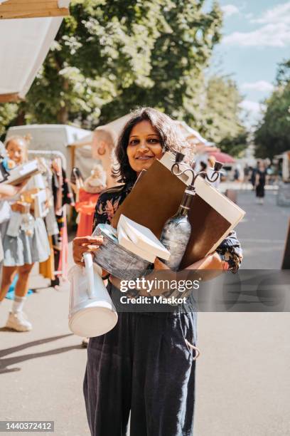 portrait of smiling woman holding various merchandise while standing at flea market - flea market stockfoto's en -beelden