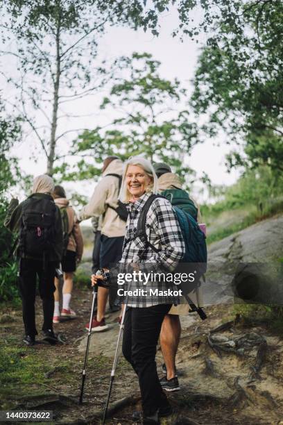 side view of smiling senior woman holding pole during hiking with friends in forest - hiking pole stock-fotos und bilder