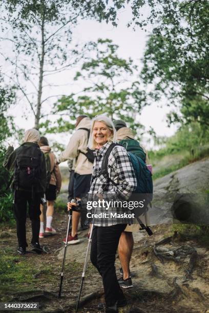 side view of smiling senior woman holding pole during hiking with friends in forest - hiking pole photos et images de collection