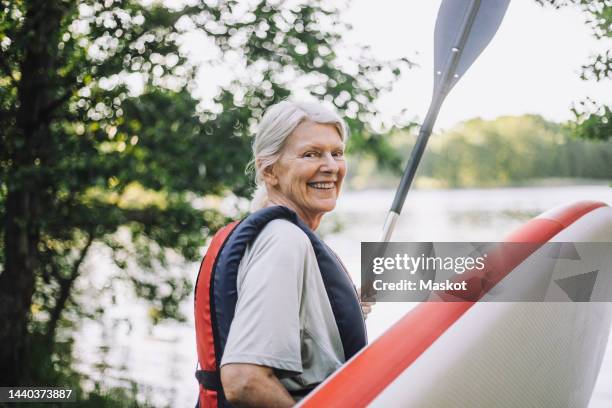 portrait of happy senior woman holding paddleboard and oar - sup stockfoto's en -beelden