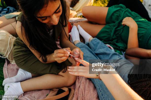smiling teenage girl applying nail polish to female friend at park - painting fingernails stock pictures, royalty-free photos & images