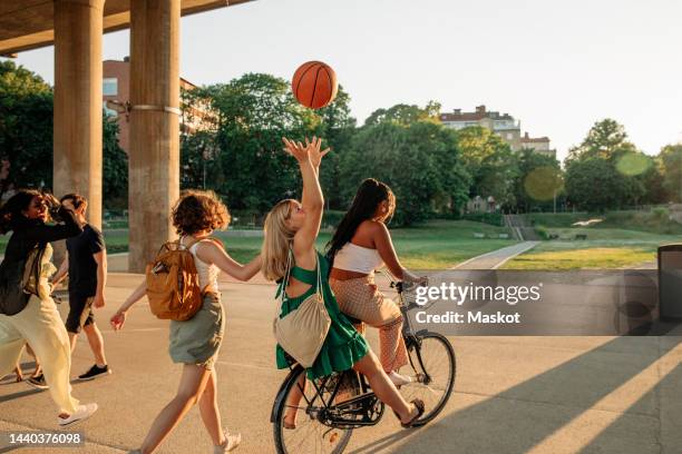 rear view of teenage girl throwing basketball while sitting with female friend riding bicycle at park - teenagers only ストックフォトと画像