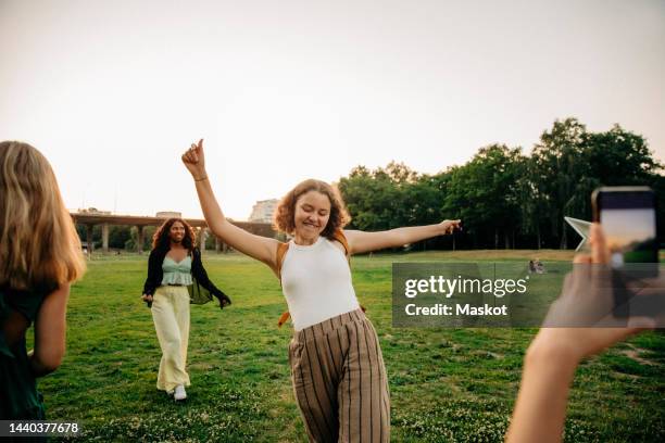 teenage girl photographing carefree female friend dancing at park during sunset - adolescenti ballano foto e immagini stock