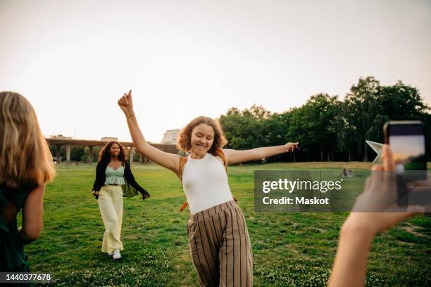 teenage girl photographing carefree female friend dancing at park during sunset - stockholm park stock pictures, royalty-free photos & images