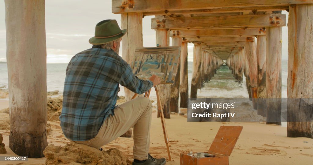 An Artist painting underneath a pier over the sea.