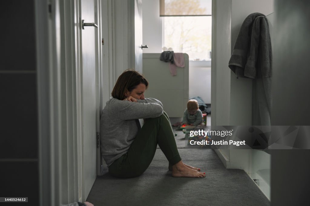 Mature mother sitting on landing floor whilst her baby plays in the background - post natal depression