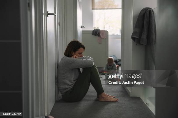 mature mother sitting on landing floor whilst her baby plays in the background - post natal depression - dark baby stock pictures, royalty-free photos & images
