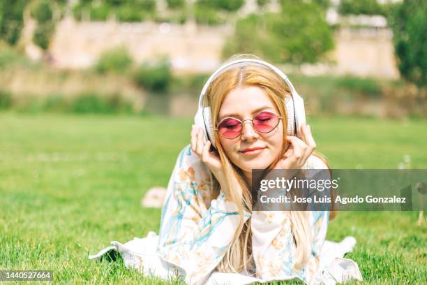 primer plano de chica joven con el pelo largo rubio, lleva puestas gafas de sol y escucha música con auriculares blancos mientras está estirada en el césped del parque de su ciudad un día soleado de primavera - primer plano stock-fotos und bilder