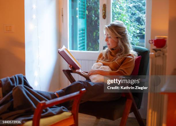 woman relaxing at home with hot water bottle, reading a book - armchair book stock pictures, royalty-free photos & images