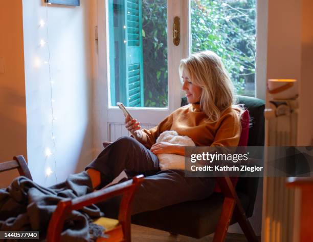 woman relaxing at home with hot water bottle, using phone - fensterladen stock-fotos und bilder