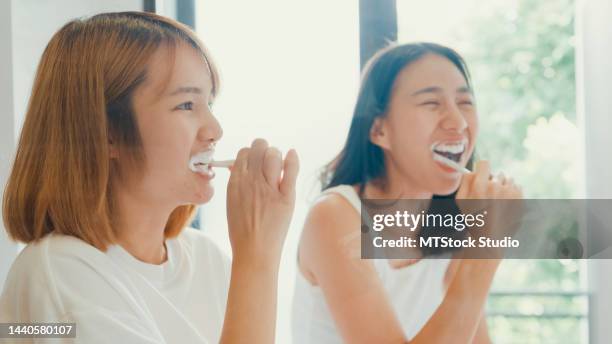 young beautiful asian lgbtq couple ladies in bathroom. happy girls having fun use toothbrush brush teeth in front of mirror together in lavatory at home. - woman in bathroom stockfoto's en -beelden