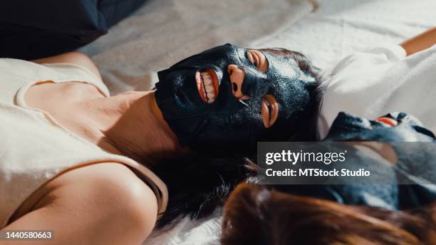 close-up young asian lesbian lgbt sweet couple ladies in bedroom. happy girls use daily face black charcoal mask on face lay on bed together at home. - antioxidants skin stock pictures, royalty-free photos & images