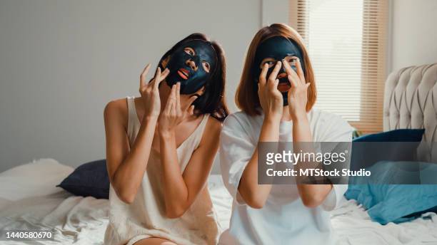 junge, schöne asiatische lesbische lgbt-paardamen im schlafzimmer. glückliche mädchen verwenden täglich gesicht schwarze holzkohle maske auf gesicht sitzen auf dem bett zusammen zu hause. - mask culture stock-fotos und bilder