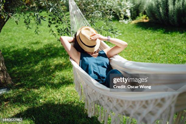 young asian woman covering her face with straw hat while taking an afternoon nap on hammock on a sunny day - hammock stockfoto's en -beelden