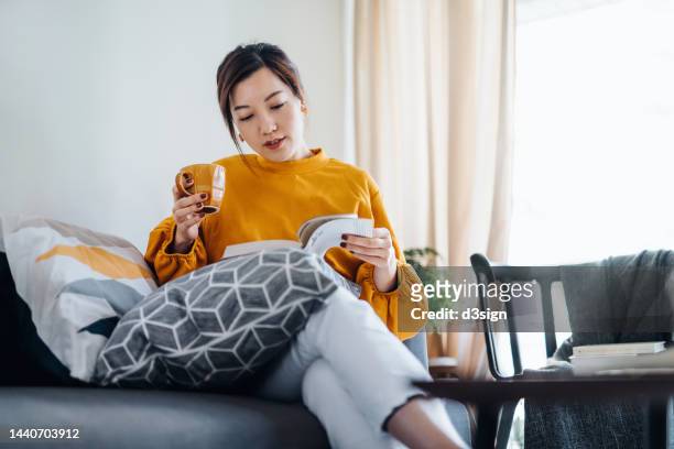 beautiful young asian woman reading a book while drinking a cup of coffee. enjoying a quiet time and relaxing environment at cozy home. digital detox time - enjoying coffee cafe morning light stock pictures, royalty-free photos & images
