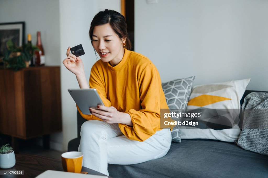 Beautiful smiling young Asian woman with credit card managing personal banking and finance with online banking mobile app on digital tablet at cozy home. Wealth management. Digital banking habits. Smart banking with technology