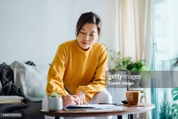 young asian woman holding a pen and signing paperwork in the living room at home. deal concept - llenar el formulario fotografías e imágenes de stock