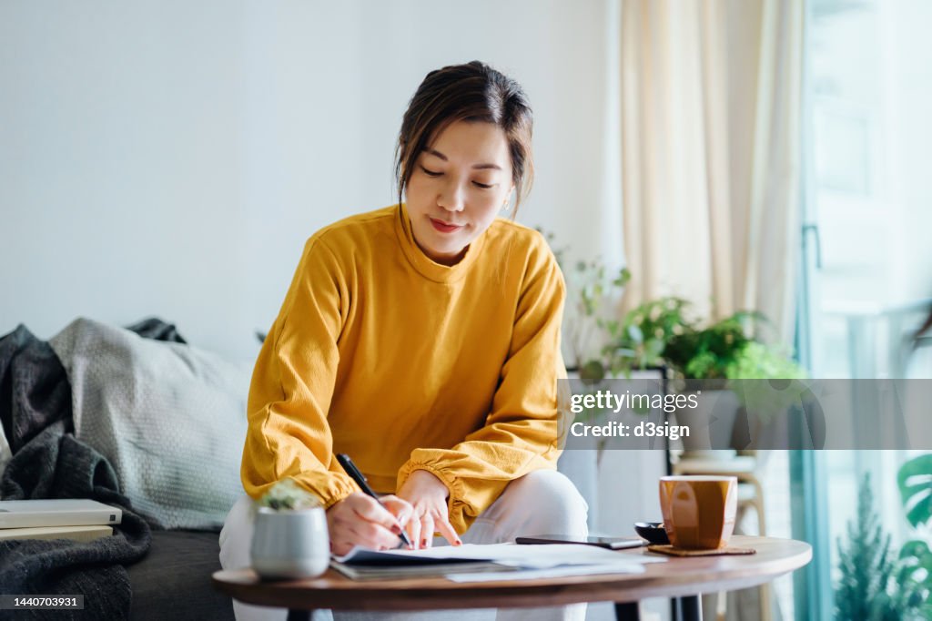 Young Asian woman holding a pen and signing paperwork in the living room at home. Deal concept