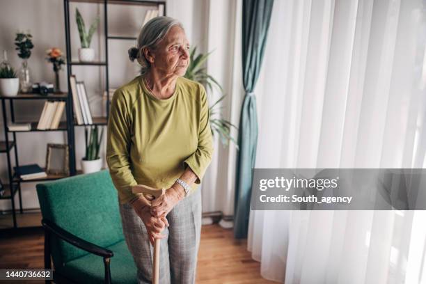 ältere frau mit gehstock schaut durch das fenster zu hause - crutch stock-fotos und bilder