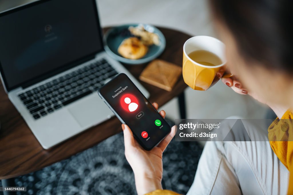 Over the shoulder view of young Asian woman receiving an incoming suspected call from unknown caller on her smartphone and rejecting the call at home. Device screen showing warning sign as detected by the network provider. Phone scam and fraud concept