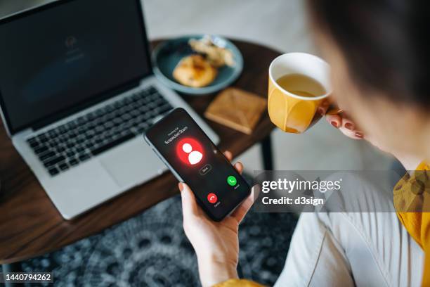 over the shoulder view of young asian woman receiving an incoming suspected call from unknown caller on her smartphone and rejecting the call at home. device screen showing warning sign as detected by the network provider. phone scam and fraud concept - alertness 個照片及圖片檔