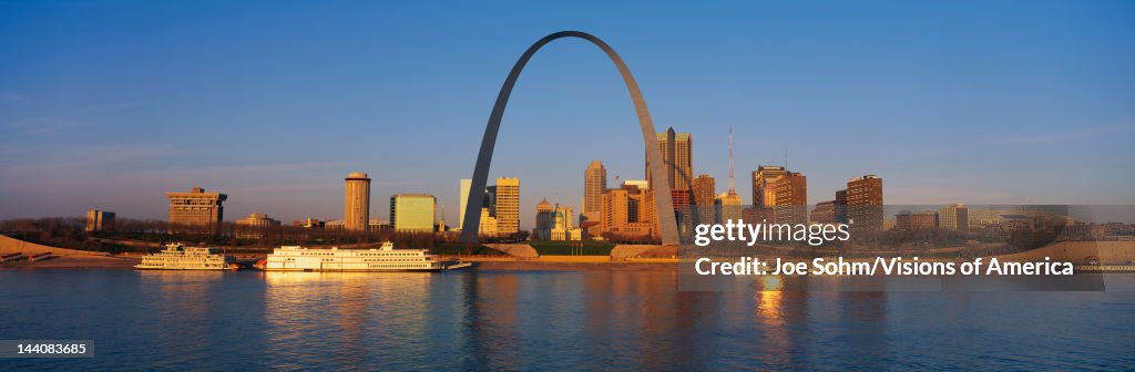 This is the skyline at sunrise. It is situated along the Mississippi River. There are riverboats on the water with the St. Louis Arch in clear view.