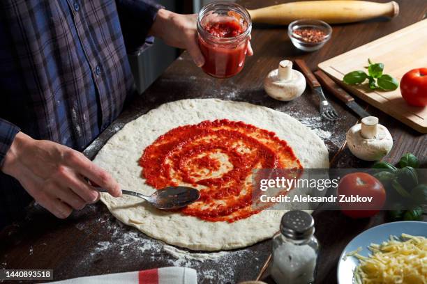 cooking italian vegetarian pizza with vegetables and mushrooms at home, pizzeria or cafe. a woman baker, pastry chef or cook spreads ketchup with a spoon on the dough, in a bakery. the concept of food. step-by-step instructions, do it yourself. step 1. - making pizza stock pictures, royalty-free photos & images