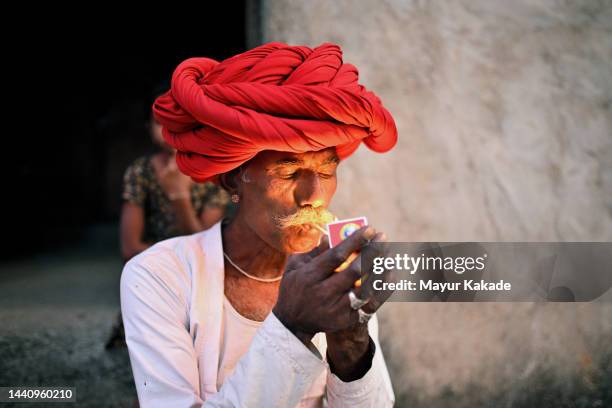 senior rabari man lighting a beedi (cigarette) sitting outside house wearing a red turban - beedi stock pictures, royalty-free photos & images