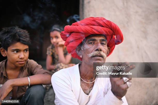 senior rabari man smoking a beedi sitting outside their house wearing a red turban looking at the camera - beedi stock pictures, royalty-free photos & images