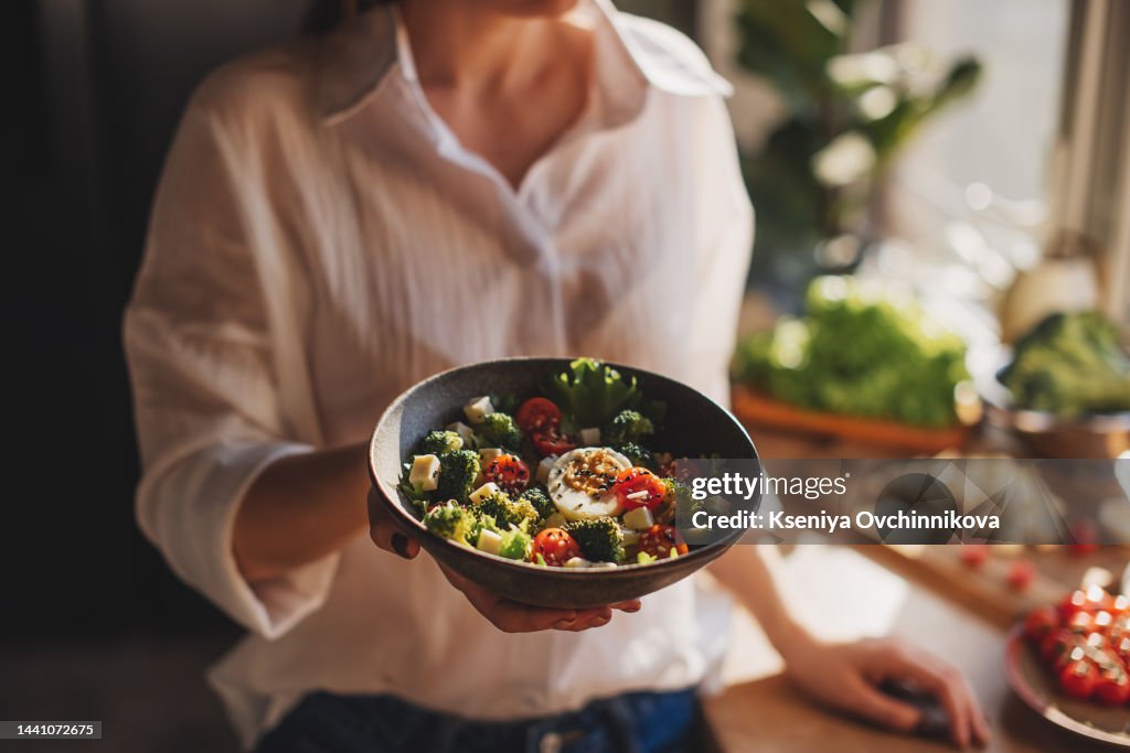 Healthy dinner or lunch. Woman in t-shirt and jeans standing and holding vegan superbowl or Buddha bowl with hummus, vegetable, salad, beans, couscous and avocado and smoothie in hands, square crop
