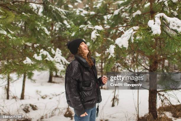 redhead curly woman in black jacket and hat in winter pine forest. pretty woman enjoying vacation. girl laughing in nature. feel happiness. hoarfrost and snow on trees. charming smile - february stockfoto's en -beelden