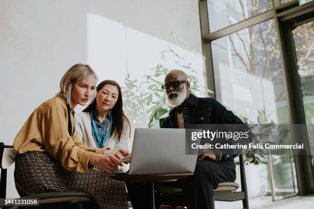 three people sit around a laptop. one woman is presenting to the group. - asesoria fotografías e imágenes de stock