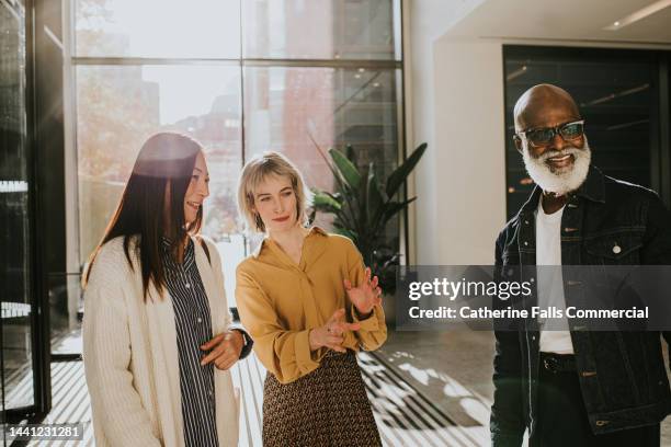 three people stand in a group in a sunny office reception. one of woman explains something to the other two participants. - wijzen handgebaar stockfoto's en -beelden