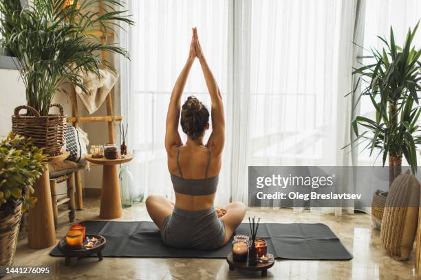 view from behind of woman meditating and practicing yoga alone at home in morning - adulto de mediana edad fotografías e imágenes de stock