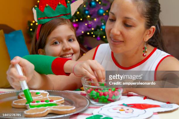 close portrait of latin family: young latin woman and a young latin girl having fun while decorating a christmas' gingerbread man for christmas night inside in a latin home in latin america, wearing christmas shirts and hat, on back, the christmas tree. - ecuador family stock pictures, royalty-free photos & images