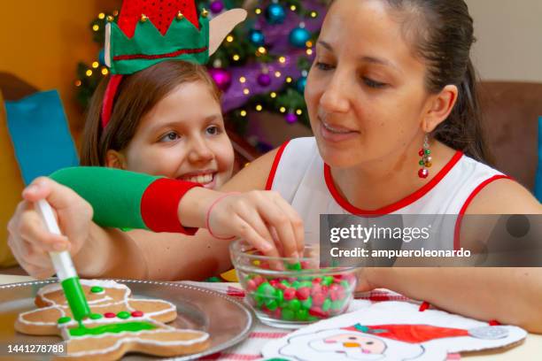 close portrait of latin family: young latin woman and a young latin girl having fun while decorating a christmas' gingerbread man for christmas night inside in a latin home in latin america, wearing christmas shirts and hat, on back, the christmas tree. - ecuador family stock pictures, royalty-free photos & images