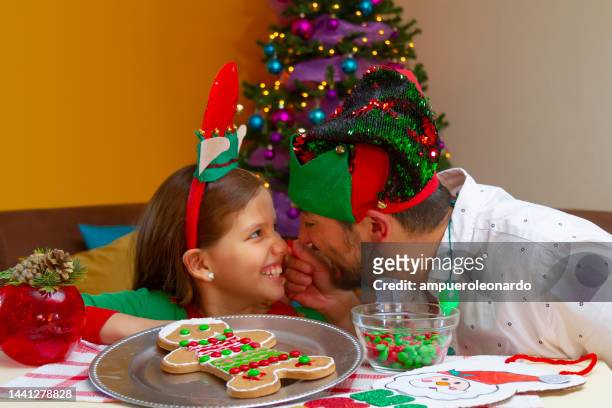 close portrait of young latin girl having fun with her dad while decorating a christmas' gingerbread man for christmas night inside in a latin home in latin america, wearing christmas shirt and hat, on back, the christmas tree. - ecuador family stock pictures, royalty-free photos & images