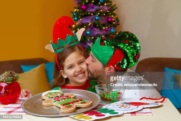 close portrait of young latin girl having fun while decorating a christmas' gingerbread man for christmas night inside in a latin home in latin america, wearing christmas shirt and hat, on back, the christmas tree. - ecuador family stock pictures, royalty-free photos & images
