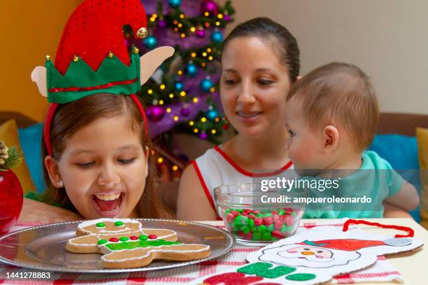 close portrait of latin family, having fun while decorating a christmas' gingerbread man for christmas night inside in a latin home in latin america, wearing christmas shirts and hat, on back, the christmas tree. - ecuador family stock pictures, royalty-free photos & images