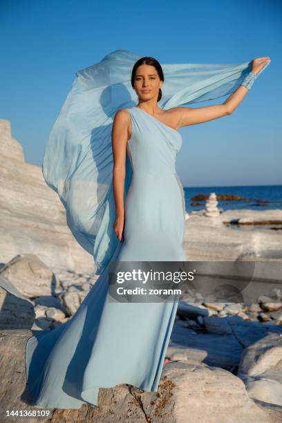 beautiful young fashion model in long light blue sleeveless dress playing with silky gown on the rocks by the sea - woman long dress beach stock pictures, royalty-free photos & images